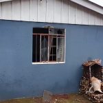 Casa azul com uma janela quebrada, pedaços de vidro no chão e ao lado, um altar com um vaso caido.