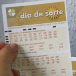 Mão segurando volante do Dia de Sorte 1224 em frente à parede branca.