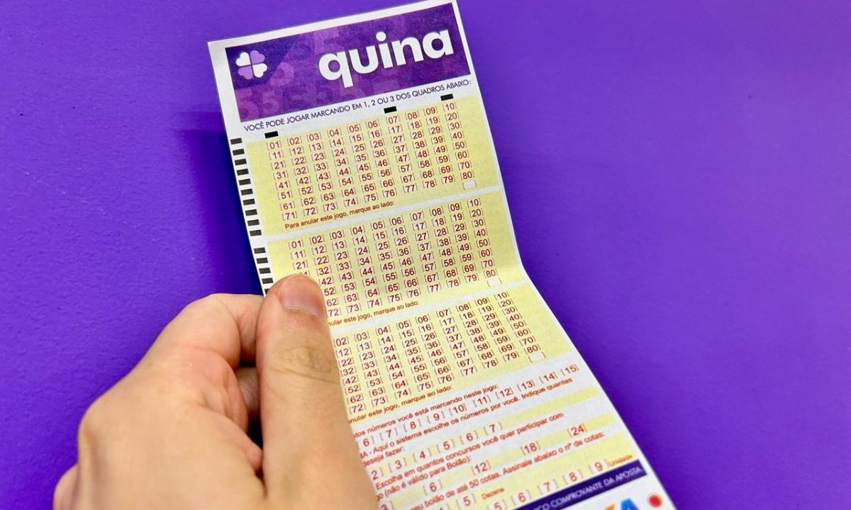 Mão segurando volante da Quina 7047 em frente à parede roxa.