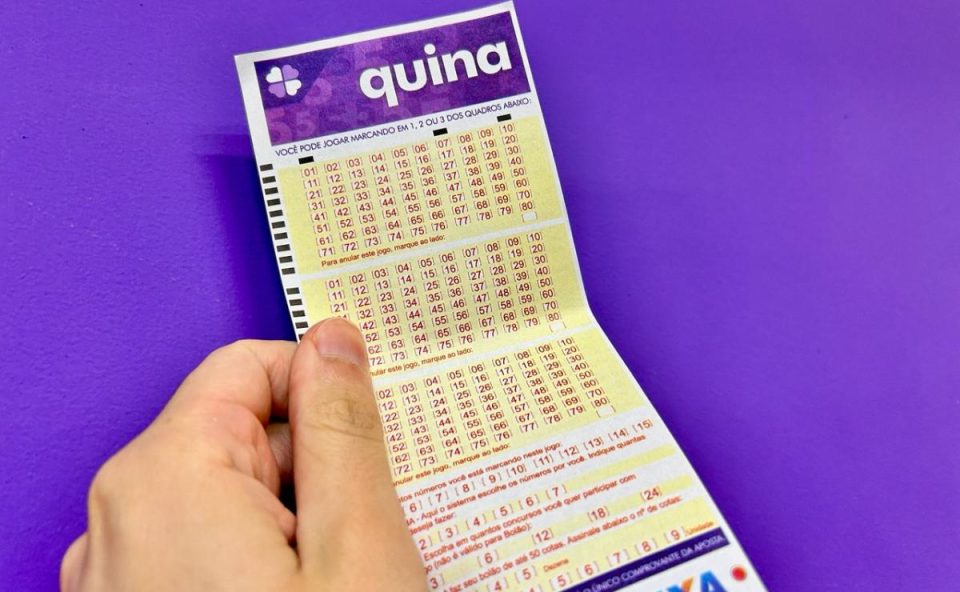 Mão segurando volante da Quina 7047 em frente à parede roxa.