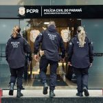foto mostra o arsenal apreendido em piraquara. suspeito foi preso. foto mostra armas em cima de uma mesa. foto mostra três policiais civis de costas.