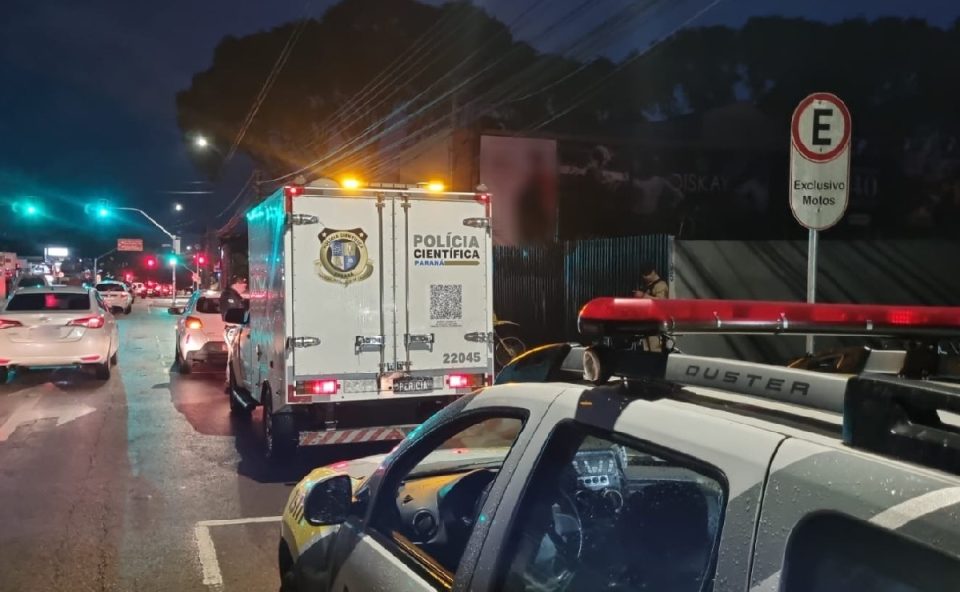 Foto registrada durante o amanhecer viaturas da Polícia Militar e da Polícia Científica em uma rua de Curitiba, com movimentação intensa. Viaturas atendem ocorrência em que suspeito morreu em confronto