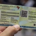 Uma mão segurando o modelo impresso em papel da Carteira de Identidade Nacional (CIN), exibindo o layout com QR Code central, foto fictícia de ilustração e campos de dados pessoais, destacando a nova carteira de identidade.